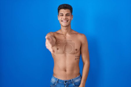 Foto de Joven hombre hispano de pie sin camisa sobre fondo azul sonriente amistoso ofreciendo apretón de manos como saludo y bienvenida. negocios exitosos. - Imagen libre de derechos