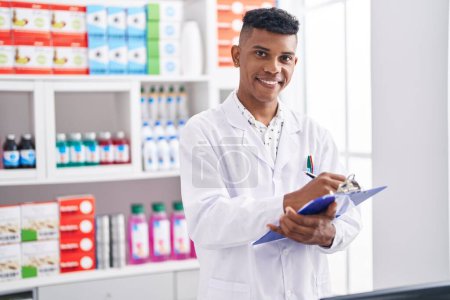 Foto de Joven farmacéutico latino sonriendo confiado informe de escritura en la farmacia - Imagen libre de derechos
