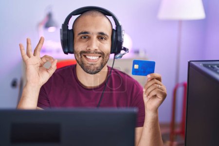 Foto de Hombre hispano con barba haciendo compras en línea con computadora y tarjeta de crédito haciendo signo ok con los dedos, sonriendo gesto amistoso excelente símbolo - Imagen libre de derechos