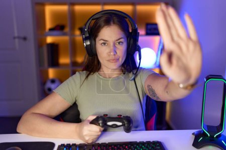 Téléchargez les photos : Belle femme brune jouant à des jeux vidéo portant des écouteurs faisant arrêter de chanter avec la paume de la main. expression d'avertissement avec geste négatif et sérieux sur le visage. - en image libre de droit