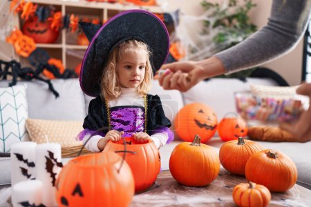 Foto de Adorable chica rubia con disfraz de bruja recibiendo dulces en la cesta de calabaza en casa - Imagen libre de derechos