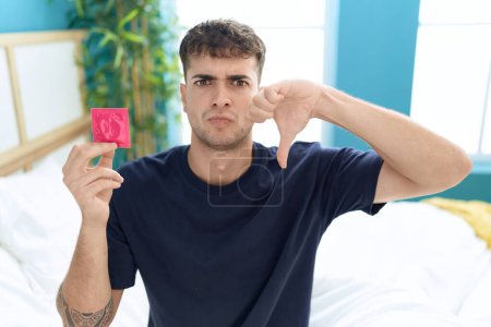 Téléchargez les photos : Jeune homme hispanique assis sur le lit tenant un préservatif avec un visage en colère, signe négatif montrant aversion avec les pouces vers le bas, concept de rejet - en image libre de droit