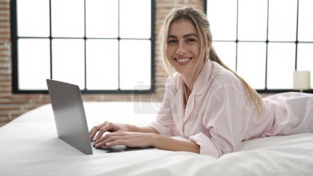 Téléchargez les photos : Jeune femme blonde utilisant un ordinateur portable couché sur le lit dans la chambre - en image libre de droit