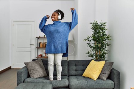 Téléchargez les photos : Femme afro-américaine écoutant de la musique danser sur le canapé à la maison - en image libre de droit