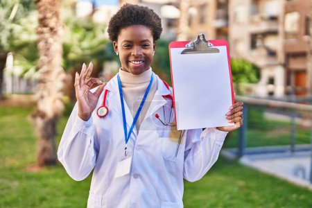 Téléchargez les photos : Afro-américaine femme portant médecin stéthoscope tenant presse-papiers faire ok signe avec les doigts, sourire geste amical excellent symbole - en image libre de droit