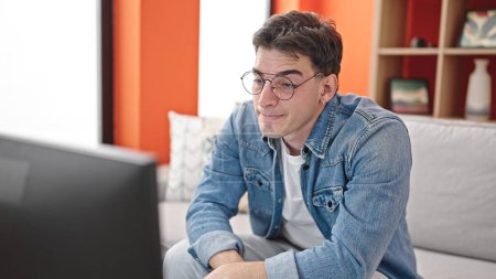 Téléchargez les photos : Jeune homme hispanique regardant la télévision portant des lunettes pour un problème de vision à la maison - en image libre de droit
