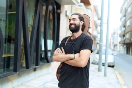 Téléchargez les photos : Young hispanic man musician holding guitar case standing with arms crossed gesture at street - en image libre de droit