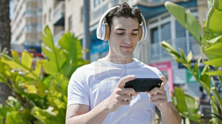 Téléchargez les photos : Jeune homme hispanique souriant confiant regarder la vidéo sur smartphone à la rue - en image libre de droit