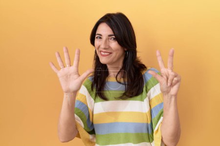 Photo for Middle age hispanic woman standing over yellow background showing and pointing up with fingers number eight while smiling confident and happy. - Royalty Free Image
