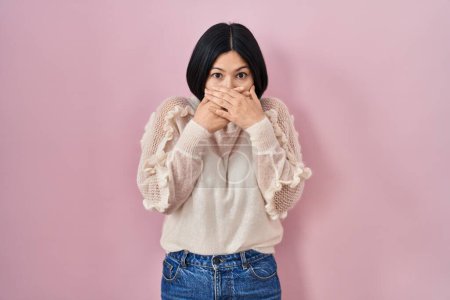 Foto de Joven mujer asiática de pie sobre fondo rosa impactado cubriendo la boca con las manos por error. concepto secreto. - Imagen libre de derechos