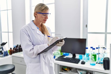 Téléchargez les photos : Jeune femme blonde portant l'uniforme de scientifique écrivant sur le presse-papiers au laboratoire - en image libre de droit