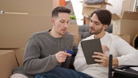 Téléchargez les photos : Deux hommes couple shopping avec pavé tactile et carte de crédit assis sur le sol à la nouvelle maison - en image libre de droit