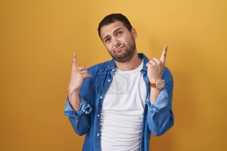 Photo for Hispanic man standing over yellow background pointing up looking sad and upset, indicating direction with fingers, unhappy and depressed. - Royalty Free Image