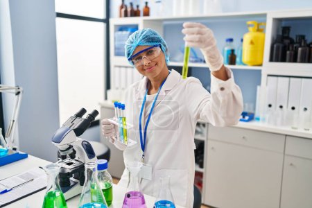 Foto de Mujer latina joven vistiendo uniforme científico sosteniendo tubo de ensayo en laboratorio - Imagen libre de derechos