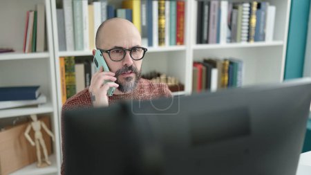 Photo for Young bald man student using computer talking on smartphone at university classroom - Royalty Free Image