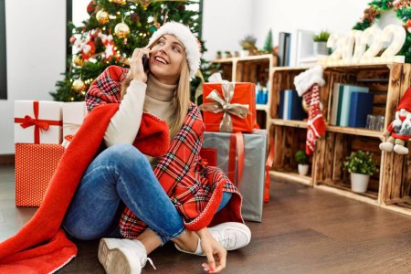 Photo for Young blonde woman talking on the smartphone sitting by christmas tree at home - Royalty Free Image