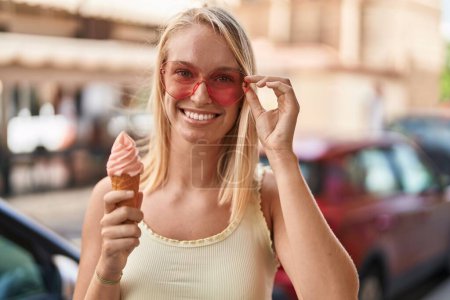 Photo for Young blonde woman wearing heart sunglasses eating ice cream at street - Royalty Free Image
