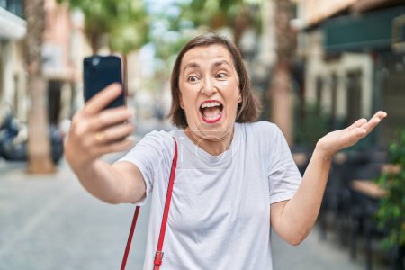 Téléchargez les photos : Moyen Age femme hispanique faire appel vidéo avec smartphone célébrer la réalisation avec sourire heureux et expression gagnante avec la main levée - en image libre de droit