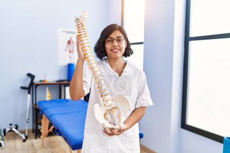 Foto de Mujer latina joven con uniforme de fisioterapeuta que sostiene el modelo anatómico de columna vertebral en la clínica de fisioterapia - Imagen libre de derechos
