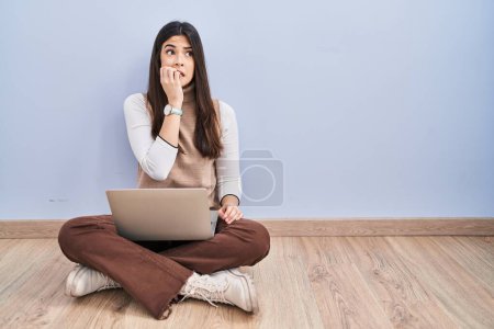Foto de Mujer morena joven trabajando con computadora portátil sentada en el suelo mirando estresada y nerviosa con las manos en la boca mordiendo clavos. problema de ansiedad. - Imagen libre de derechos