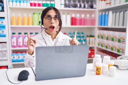 Téléchargez les photos : Jeune femme arabe travaillant à la pharmacie pharmacie en utilisant un ordinateur portable étonné et surpris de regarder vers le haut et pointant avec les doigts et les bras levés. - en image libre de droit