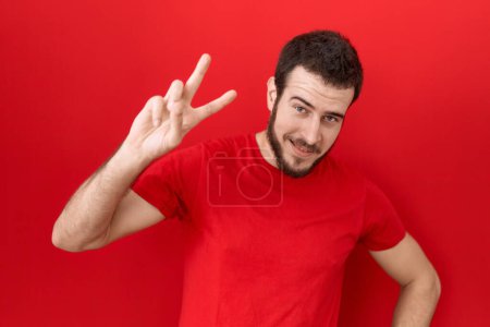 Foto de Joven hombre hispano con camiseta roja casual sonriendo mirando a la cámara mostrando los dedos haciendo señal de victoria. número dos. - Imagen libre de derechos