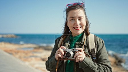 Téléchargez les photos : Femme hispanique mature avec des cheveux gris touriste portant un sac à dos tenant caméra vintage au bord de la mer - en image libre de droit