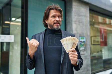 Photo for Handsome middle age man holding 100 danish krone banknotes pointing thumb up to the side smiling happy with open mouth - Royalty Free Image