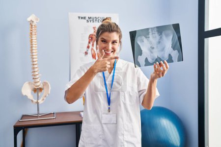 Foto de Mujer joven sosteniendo radiografía de pelvis sonriendo feliz señalando con la mano y el dedo - Imagen libre de derechos