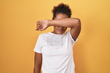 Photo for Young hispanic woman with curly hair standing over yellow background covering eyes with arm, looking serious and sad. sightless, hiding and rejection concept - Royalty Free Image