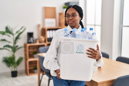 Téléchargez les photos : Femme afro-américaine travaillant au bureau tenant une bouteille en plastique pour le recyclage souriant regardant vers le côté et regardant loin en pensant. - en image libre de droit