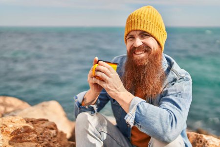 Téléchargez les photos : Young redhead man smiling confident drinking cup of coffee at seaside - en image libre de droit
