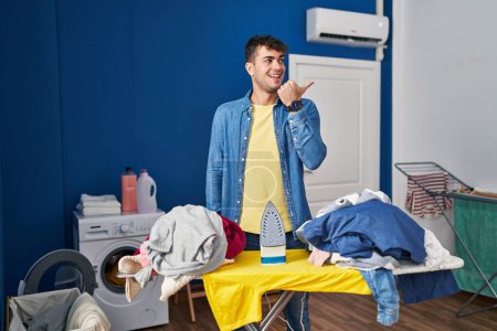 Téléchargez les photos : Jeune homme hispanique repassage vêtements à la maison pointant pouce vers le côté souriant heureux avec la bouche ouverte - en image libre de droit