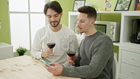 Photo for Two men couple using smartphone drinking glass of wine at dinning room - Royalty Free Image