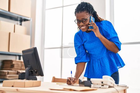 Téléchargez les photos : Africaine américaine femme commerce électronique travailleur d'affaires parler smartphone écriture sur ordinateur portable au bureau - en image libre de droit