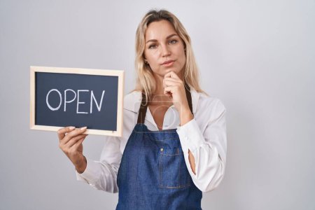 Photo for Young blonde woman wearing apron holding blackboard with open word serious face thinking about question with hand on chin, thoughtful about confusing idea - Royalty Free Image