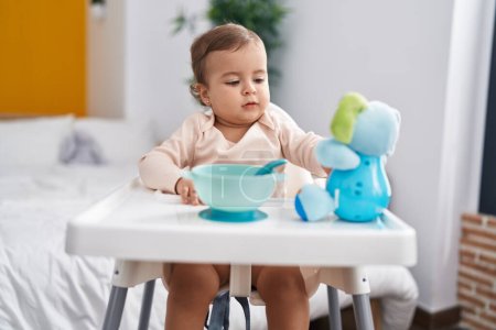 Foto de Adorable bebé hispano sentado en una silla alta jugando con un juguete de elefante en el dormitorio - Imagen libre de derechos