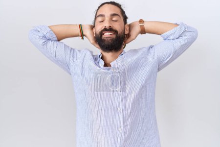 Téléchargez les photos : Homme hispanique avec barbe vêtu d'une chemise décontractée relaxante et étirante, bras et mains derrière la tête et le cou souriant heureux - en image libre de droit