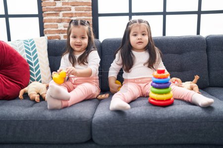Foto de Adorable gemelas jugando con aros juego sosteniendo pato juguete sentado en el sofá en casa - Imagen libre de derechos