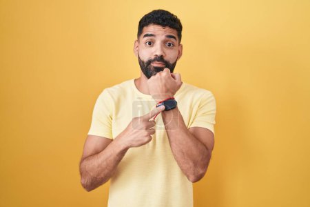 Téléchargez les photos : Homme hispanique avec barbe debout sur fond jaune à la hâte pointant vers regarder le temps, l'impatience, regardant la caméra avec une expression détendue - en image libre de droit