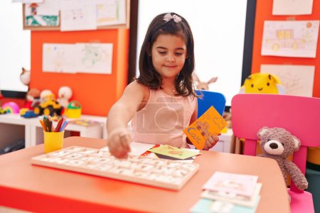 Photo for Adorable hispanic girl playing with vocabulary puzzle game sitting on table at kindergarten - Royalty Free Image