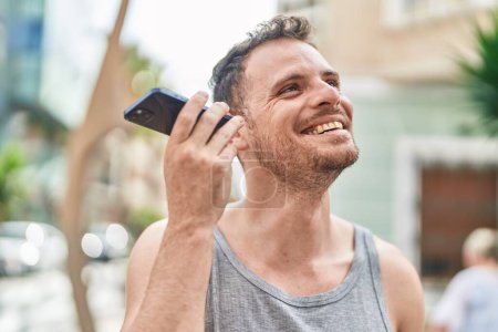 Téléchargez les photos : Jeune homme hispanique souriant confiant écoute message audio par le smartphone à la rue - en image libre de droit