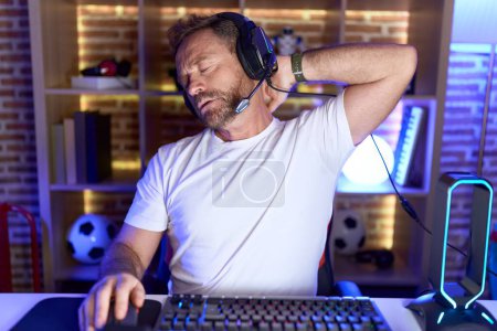 Photo for Middle age man with beard playing video games wearing headphones suffering of neck ache injury, touching neck with hand, muscular pain - Royalty Free Image