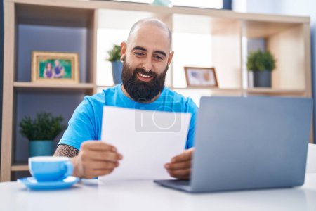 Photo for Young bald man using laptop reading paper at home - Royalty Free Image
