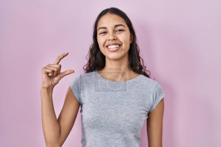 Foto de Mujer brasileña joven con camiseta casual sobre fondo rosa sonriente y gesto seguro con la mano haciendo signo de tamaño pequeño con los dedos mirando y la cámara. concepto de medida. - Imagen libre de derechos