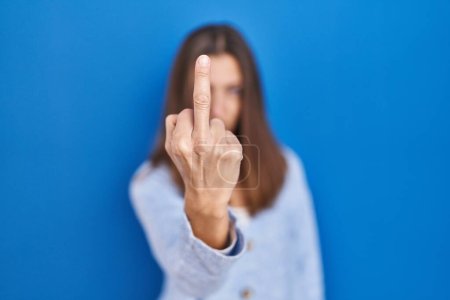 Foto de Young woman standing over blue background showing middle finger, impolite and rude fuck off expression - Imagen libre de derechos
