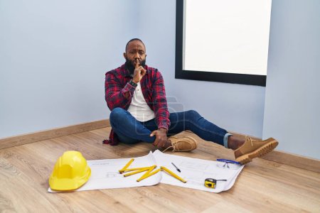 Photo for African american man sitting on the floor at new home looking at blueprints asking to be quiet with finger on lips. silence and secret concept. - Royalty Free Image