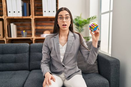Téléchargez les photos : Hispanic woman playing colorful puzzle cube intelligence game scared and amazed with open mouth for surprise, disbelief face - en image libre de droit