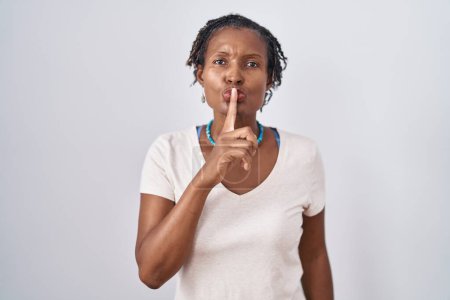 Téléchargez les photos : Femme africaine avec dreadlocks debout sur fond blanc demandant à être calme avec le doigt sur les lèvres. silence et concept secret. - en image libre de droit