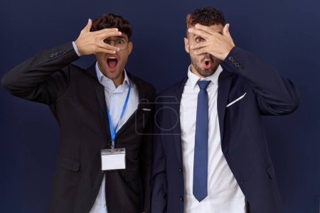 Foto de Dos hombres de negocios hispanos vistiendo ropa de negocios asomándose en shock cubriendo cara y ojos con la mano, mirando a través de los dedos con expresión avergonzada. - Imagen libre de derechos
